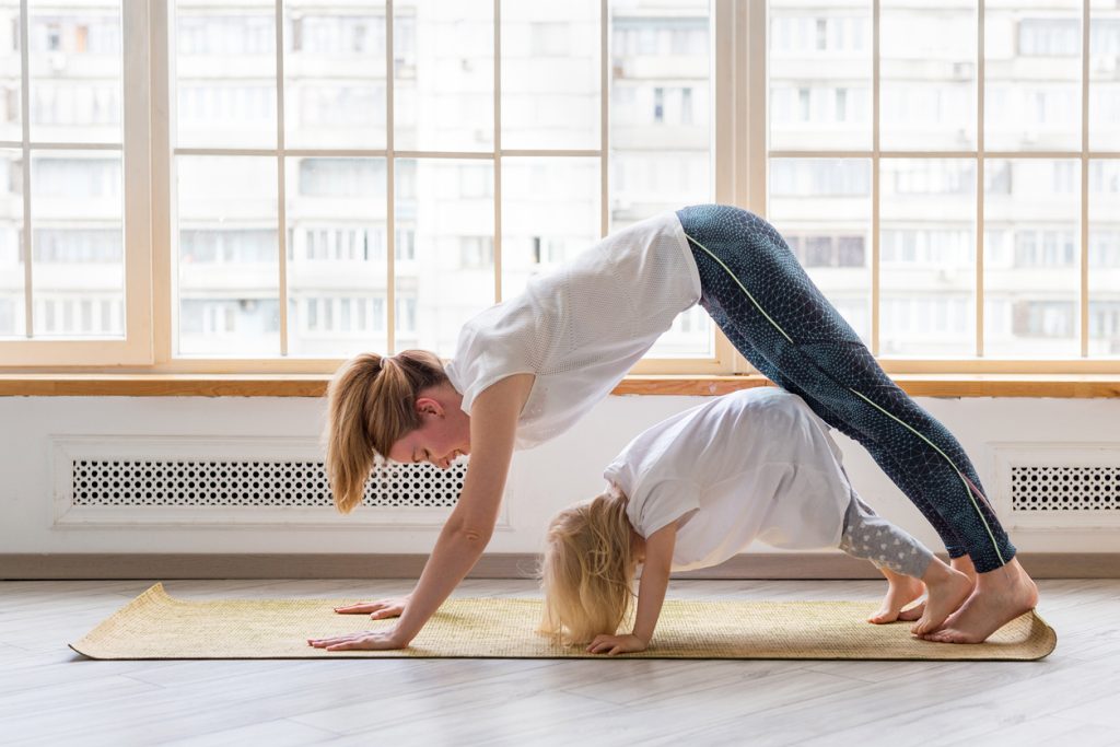 The 5 best ways to exercise as a family 1 Young Mother Doing Yoga With 3-Years Girl In Front Of Window. Downward Facing Dog Asana